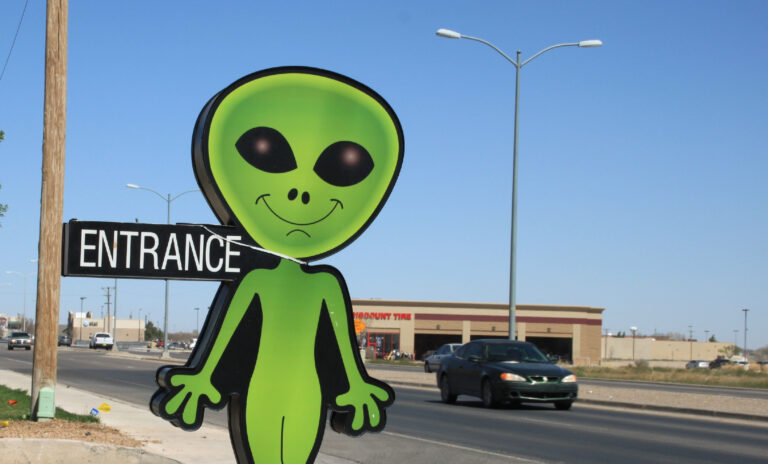 Eine lebensgroße Pappfigur eines stereotypischen grünen Außerirdischen an einer Straße, über die gerade ein Auto fährt.
