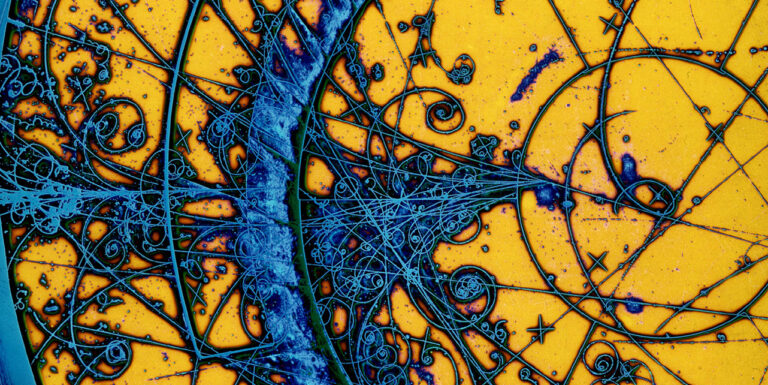 Eine kolorierte Aufnahme einer Bubble Chamber zeigt einen gelben Hintergrund mit blau eingefärbten Teilchenspuren, die teilweise gebogen und spiralförmig sind.