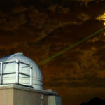 Von einer weißen Teleskopkuppel geht in einer leicht wolkigen Nacht ein grüner breiter Laserstrahl zum Mond.