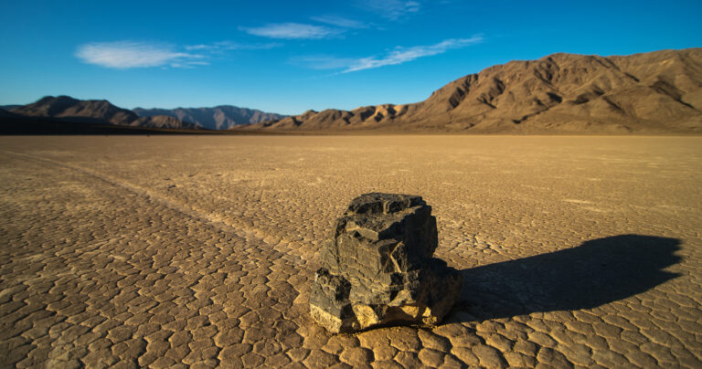 Blauer Himmel, ein dunkler Hang, davor eine beige flache Wüste: Darin liegt ein Stein, mit einer geraden Spur hinter sich.