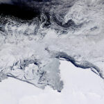 Eine Landschaft aus Satelliten-Perspektive: eine eisbedeckte Küste, eine Zone mit Eisbergen, dunkel erscheinendes Wasser