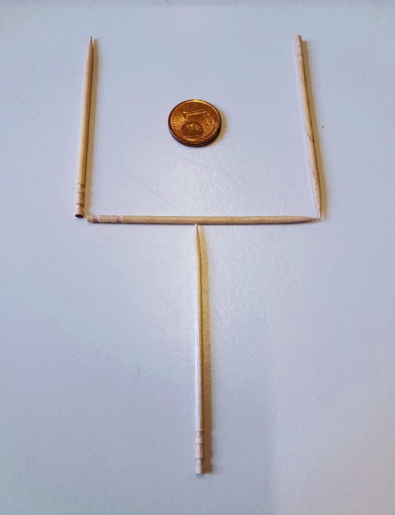 Photo of three toothpicks forming a wine glass 'cup' around a coin on a table, with a fourth below in the middle to form a 'stem'