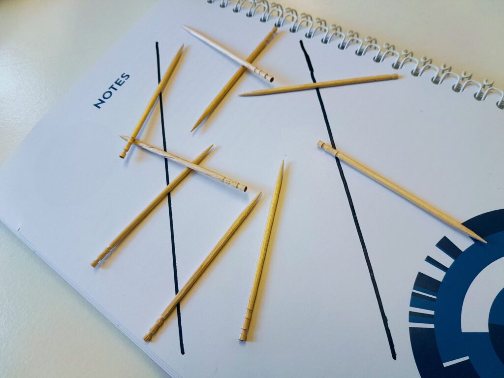 Photo of some toothpicks on a HLF branded notebook; there are two parallel lines drawn on the notebook, one toothpick length apart