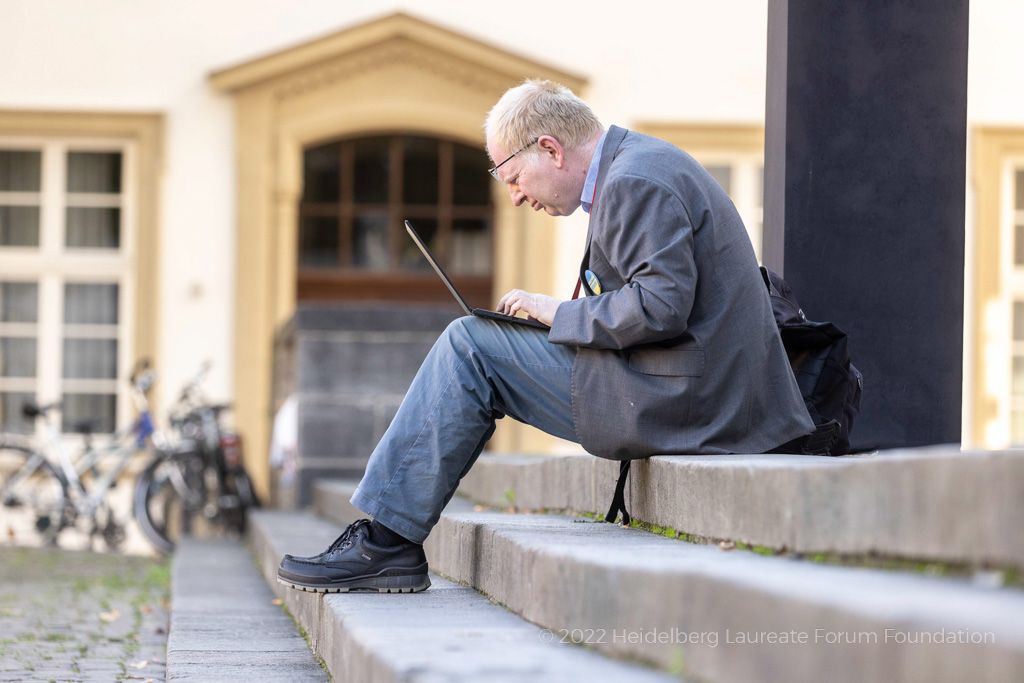 A white man with blonde hair sits on some stone steps. He is wearing a great blazer and great trousers. Some glasses rest on his head. He is typing on a laptop. In the background are some bikes. 