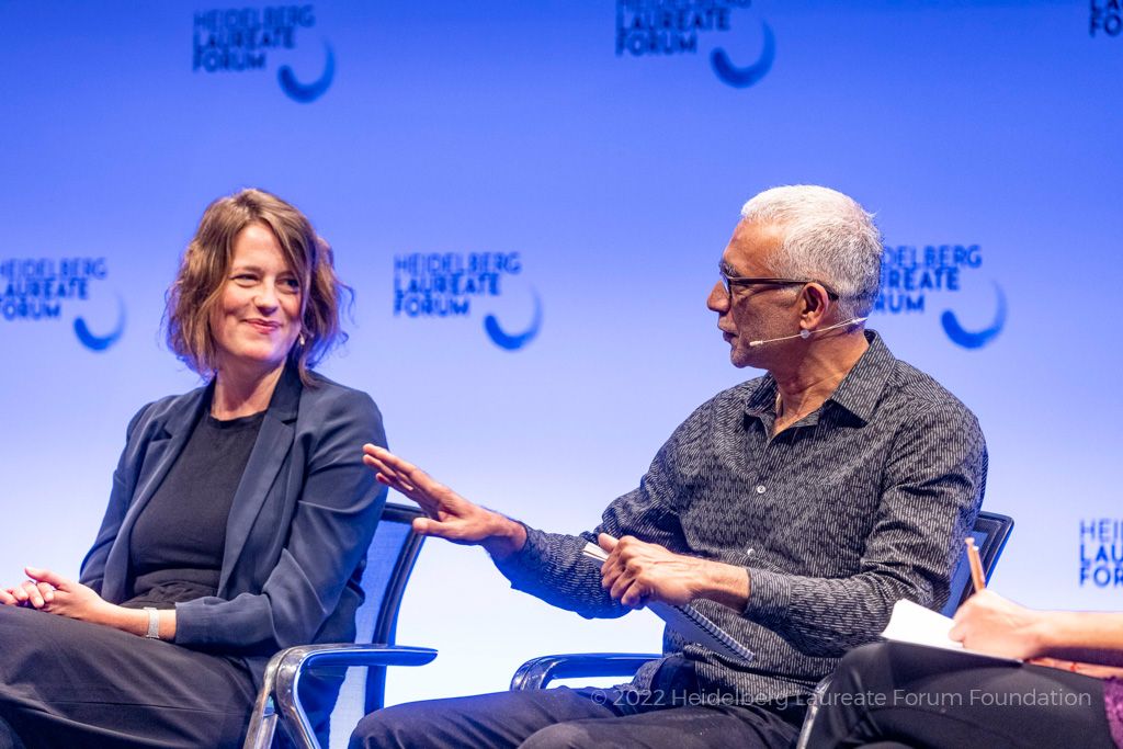 A photo of Anna Maria Hartkopf, a white woman with long wavy light-brown hair wearing a suit, and Anil Ananthaswamy, an older Indian man with grey hair and glasses wearing a grey shirt. Anil is talking animatedly.
