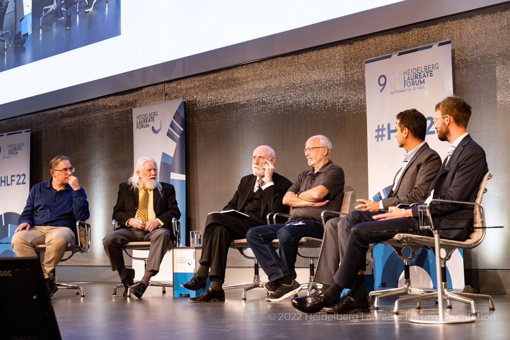 On a stage sits five white men. From left to right they are Tom Geller, Whitfield Diffie, Vinton Cerf, Avi Shamir, Vadim Lyubashevsky, and Gregor Seiler. The camera is just beyond Gregor so is looking at the panel from the right.