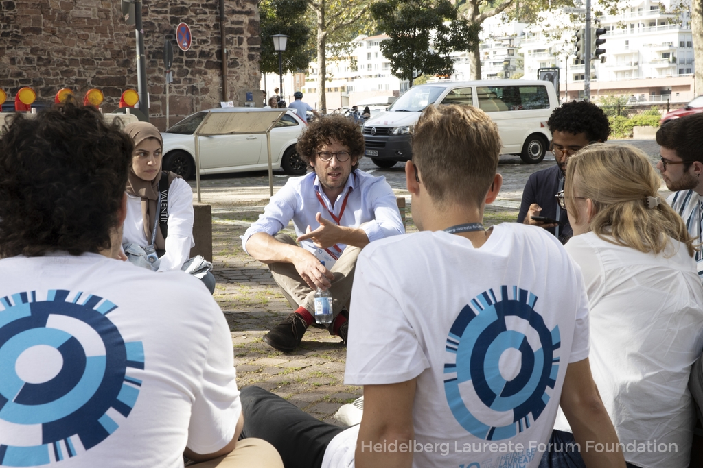 Hugo Duminil-Copin sits on the floor cross-legged talking to young researchers. The young researchers are wearing HLF branded t-shirts and sitting in a circle around him