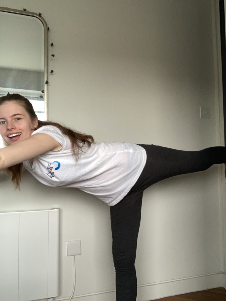 A young blonde woman is wearing a white Heidelberg Laureate Forum t-shirt and grey leggings. She is standing on her right leg, with her arms reaching forward, and her left leg horizontally out behind her. Her body and legs form a T shape. She is grinning/