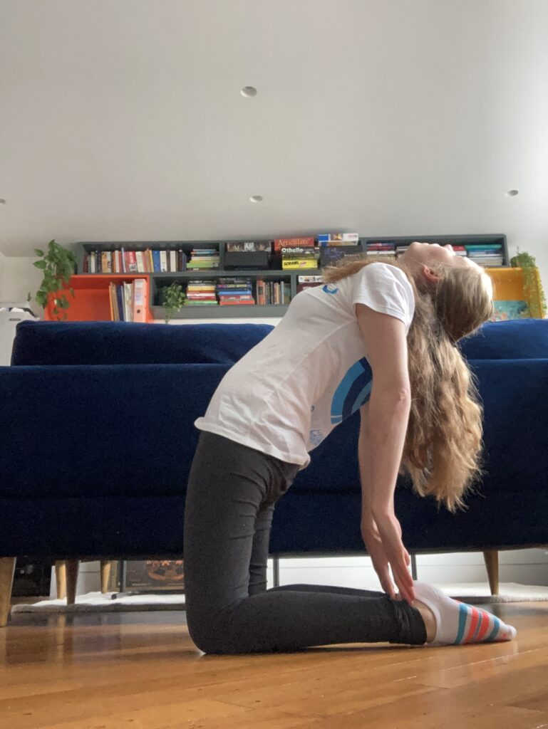 A young blonde woman is wearing a white Heidelberg Laureate Forum t-shirt and grey leggings. She is kneeling on the floor, with her knees forming a right angle. The is looking up to the sky and her hands are touching her feet.