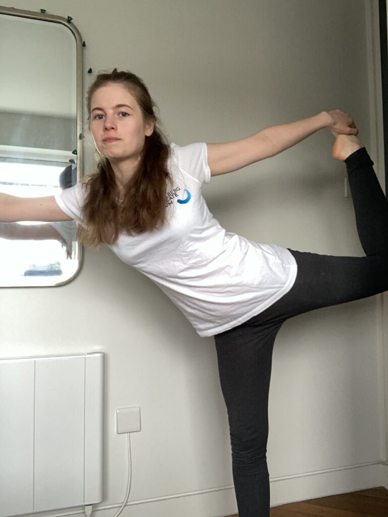 A young blonde woman is wearing a white Heidelberg Laureate Forum t-shirt and grey leggings. She is standing on her right leg and holding her left leg out behind her. Her left knee makes a right angle, and she is leaning forward slightly.