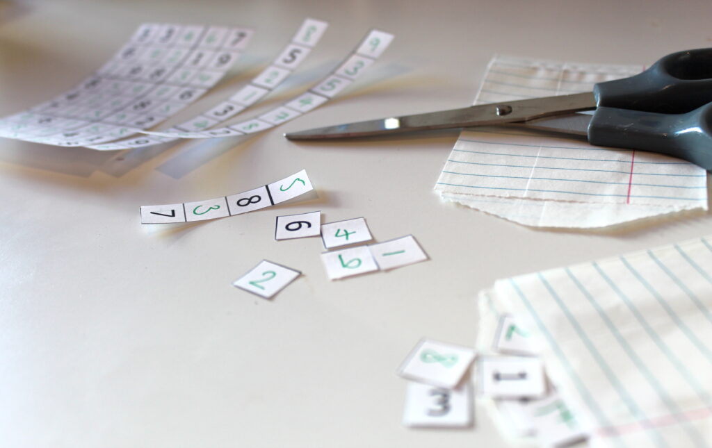 A photograph showing a partially cut up sudoku puzzle, cut into columns with a few of the columns cut into individual squares; a pair of scissors is on the table and some of the sets of squares are half-in some paper envelopes