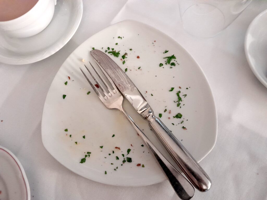 A photograph of a triangular plate with curved sides, with the remains of some breakfast and a knife and fork laid across it, on a breakfast table surrounded by other plates and cups