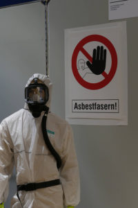 Achtung, Asbest! Ein Mensch in voller Schutzausrüstung unter einem Warnschild vor Asbest