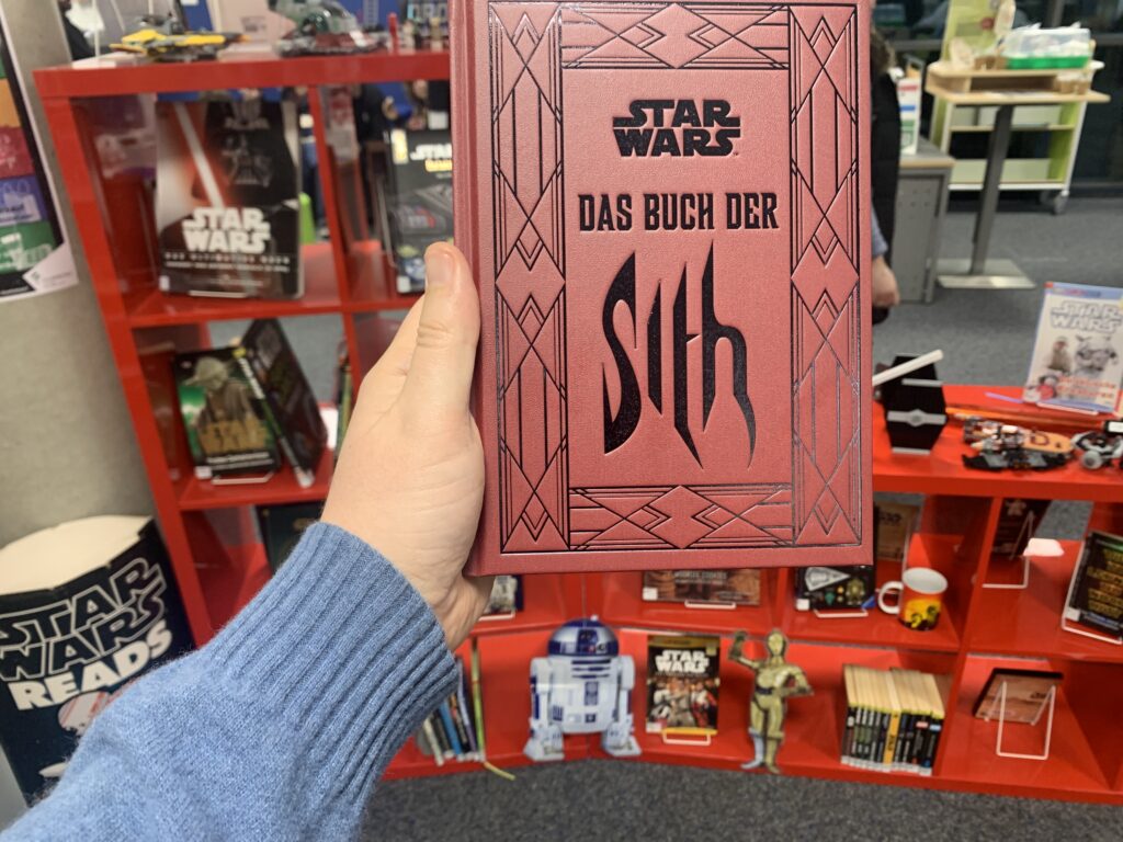 Ein Shelfie von Michael Blume feiert in der Stadtbibliothek Filderstadt "Das Buch der Sith" vor einem roten Regal voller Star Wars-Devotionalien.