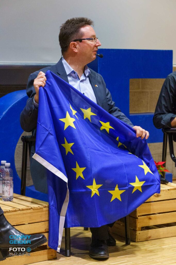 Dr. Michael Blume bei Pulse of Europa in der VHS Stuttgart sitzend, eine EU-Fahne erläuternd. Link führt zum eBooklet "A wie Aachen" zur Alphabetgeschichte Europas.
