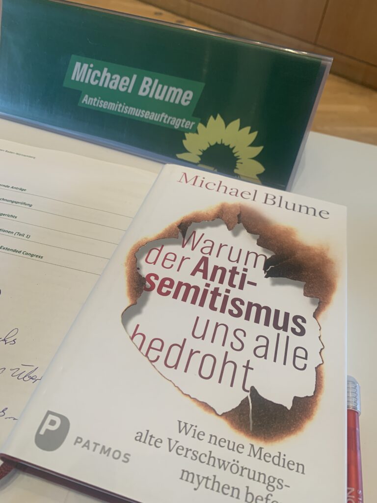 Foto vom grünen Parteitagstisch von Dr. Michael Blume zu Weingarten mit einem Namensschild, Notizen und einem Signierexemplar von "Warum der Antisemitismus uns alle bedroht".